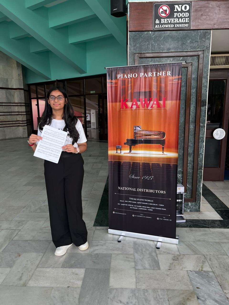 Priyal holding her sheet music next to the Kawai piano partner banner at the concert venue