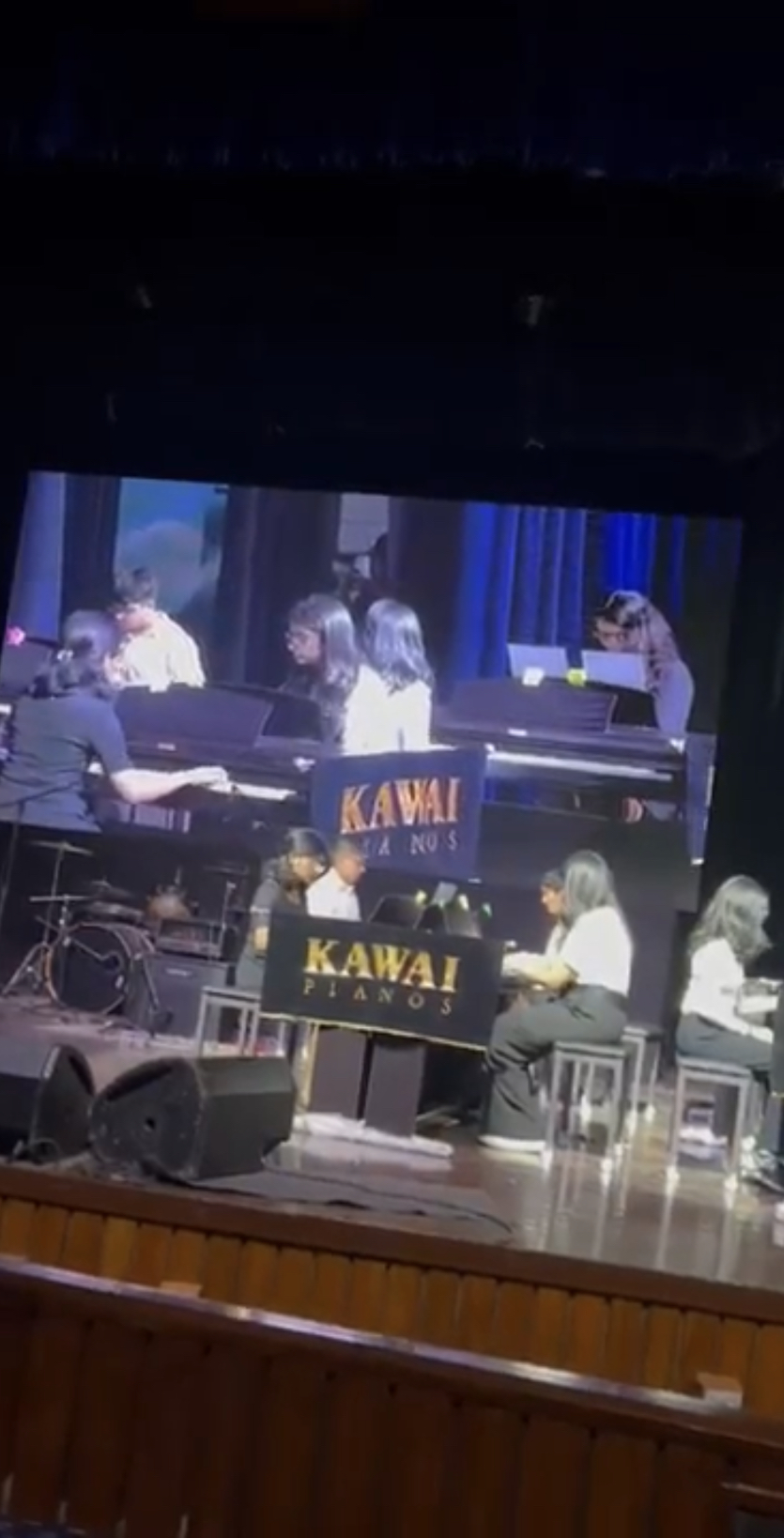 The group performing on pianos on stage at the annual concert, with the performance shown on the big screen above
