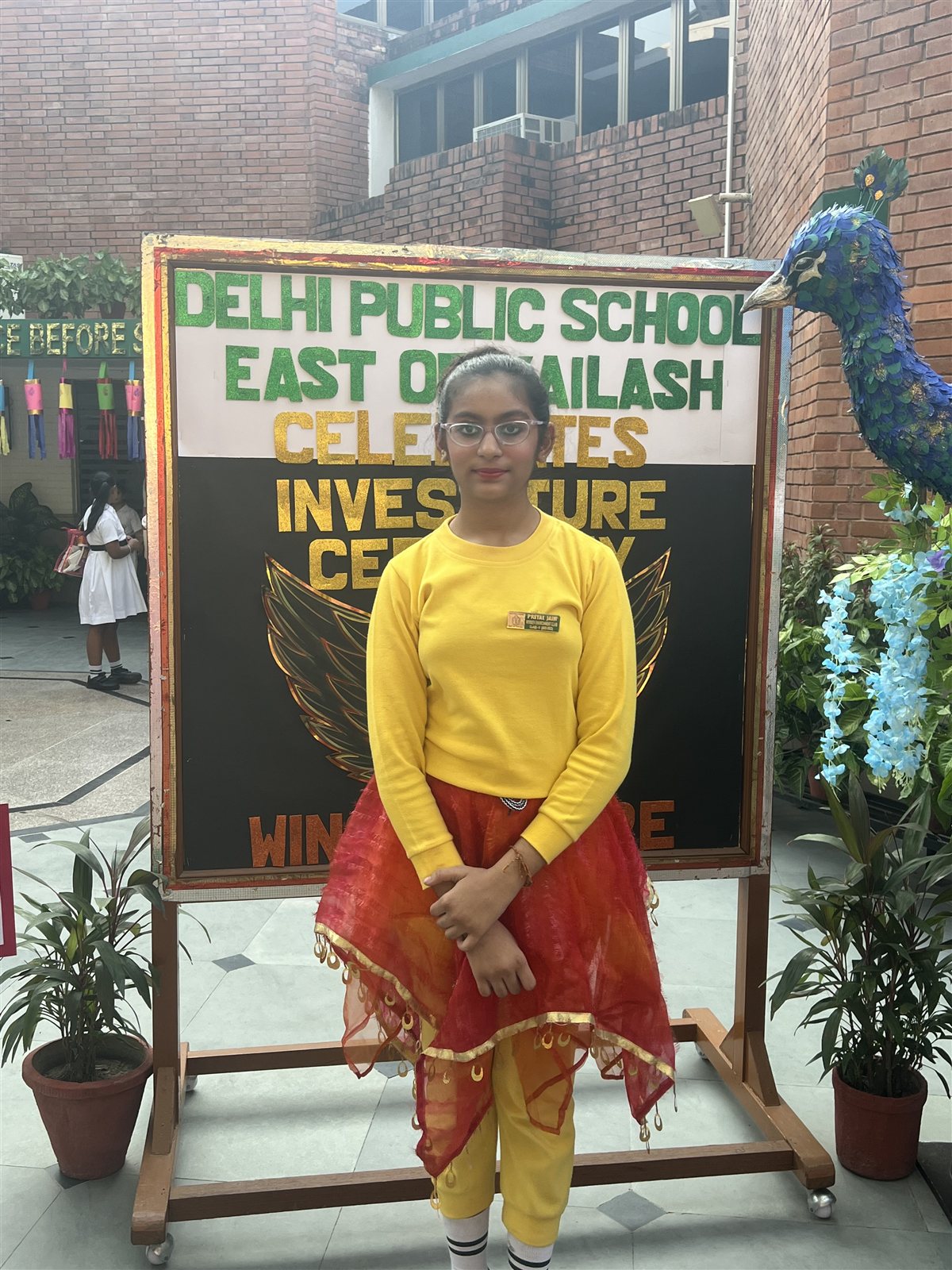 Priyal wearing her Environment Club badge in front of the Investiture Ceremony board at DPS East of Kailash